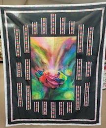 Quilt panel with a rainbow of colors coming from the center of a rose bordered by black fabric and rainbow strips of fabric.